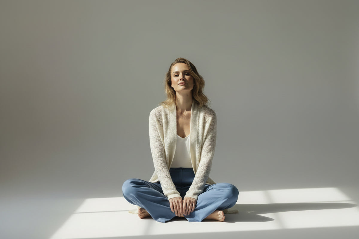A woman stands barefoot in a studio wearing a long cream textured cardigan over a white top and blue wide-leg pants against a light gray background.